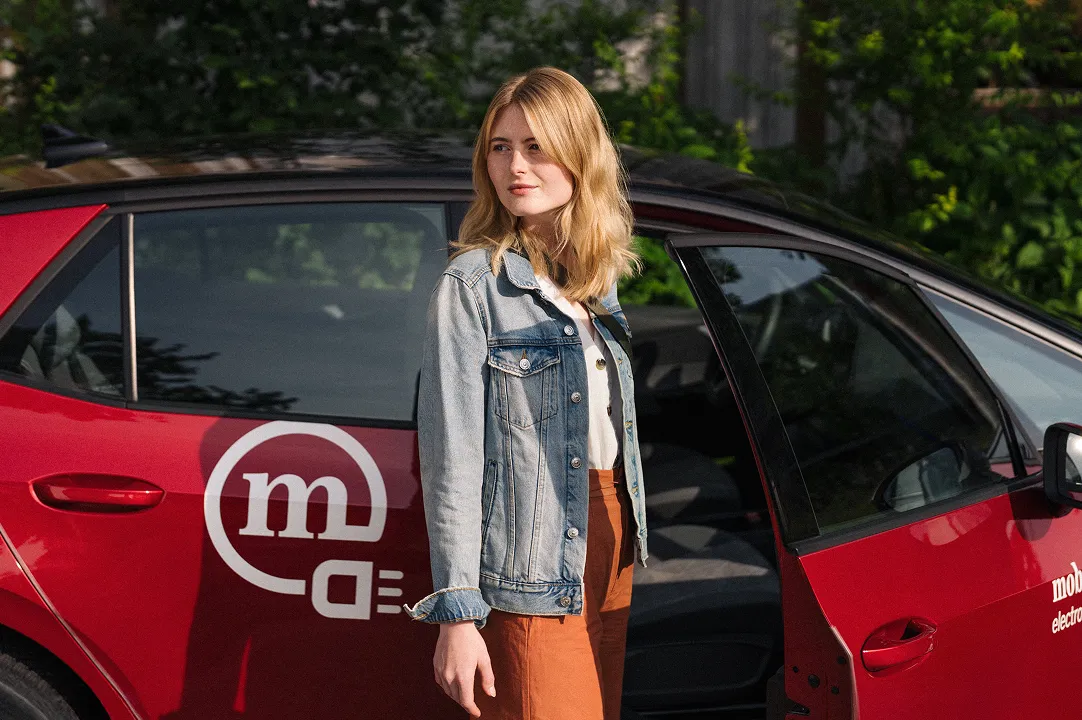 Frau steht neben rotem Mobility-Carsharing-Auto mit geöffneter Tür und nutzt geteilte Mobilität im Alltag.