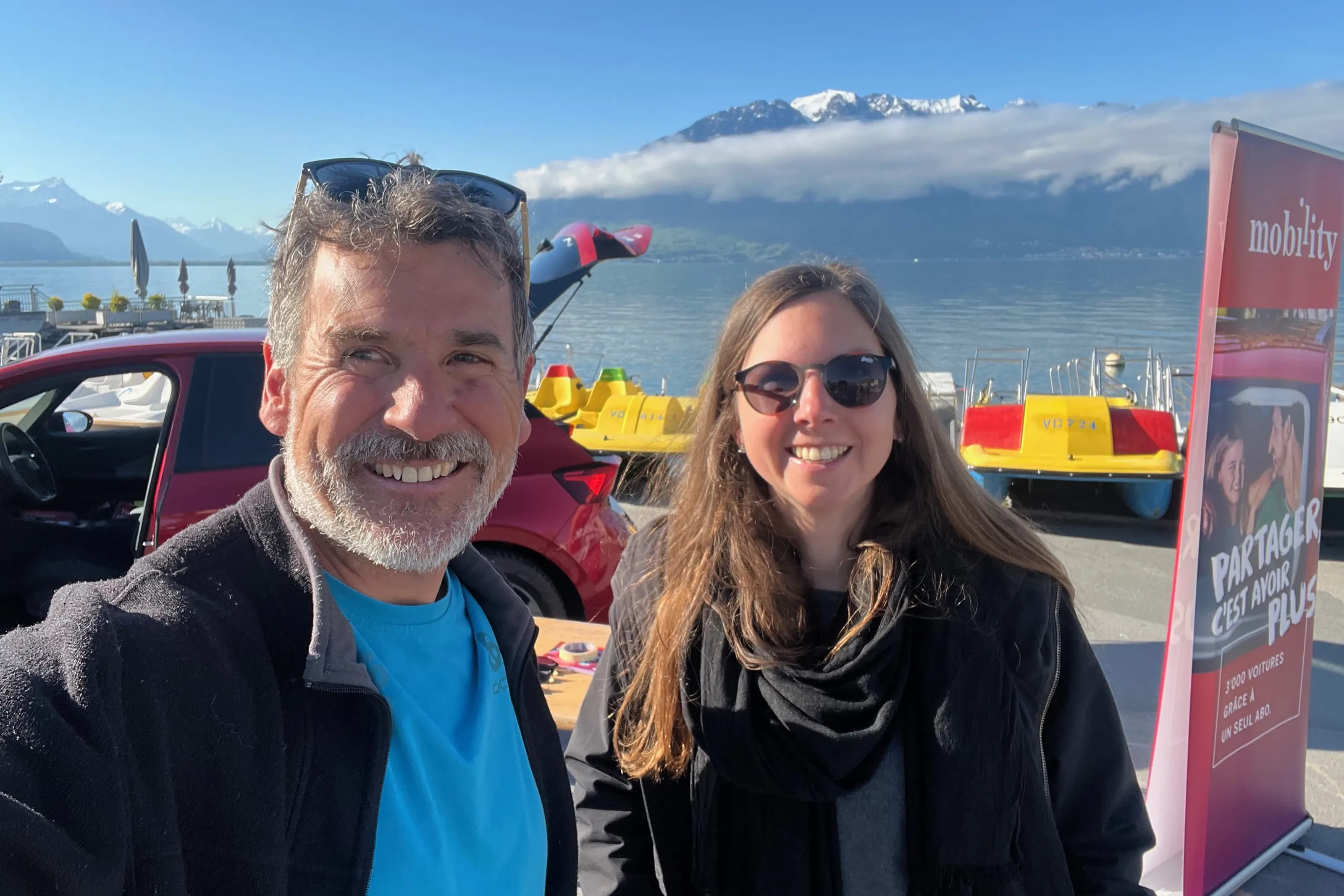 Zwei Personen machen ein Selfie am See vor Bergen neben einem roten Carsharing-Auto und Tretbooten.