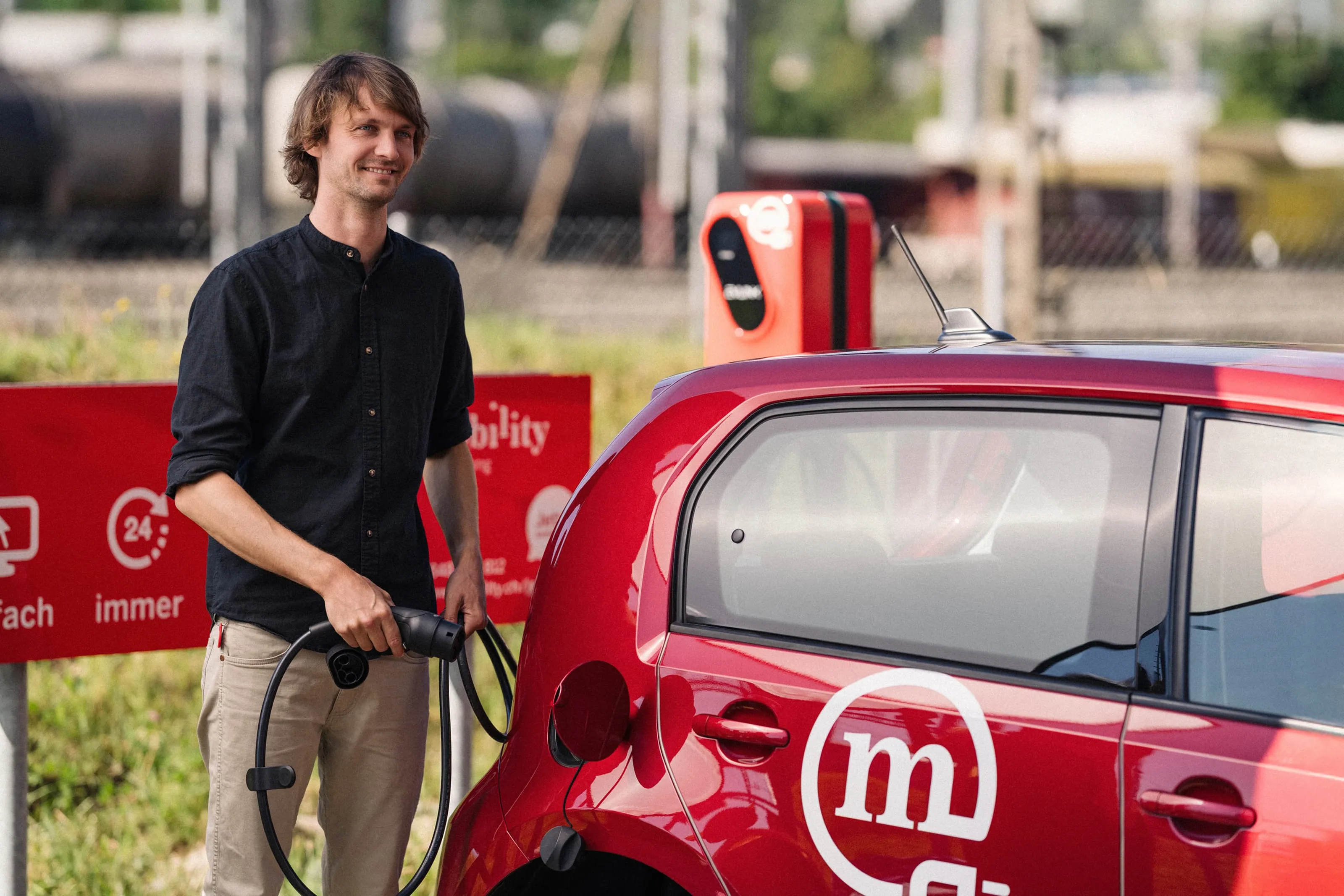 Mann lädt ein rotes Mobility-Carsharing-Elektroauto an einer öffentlichen Ladestation und lächelt.