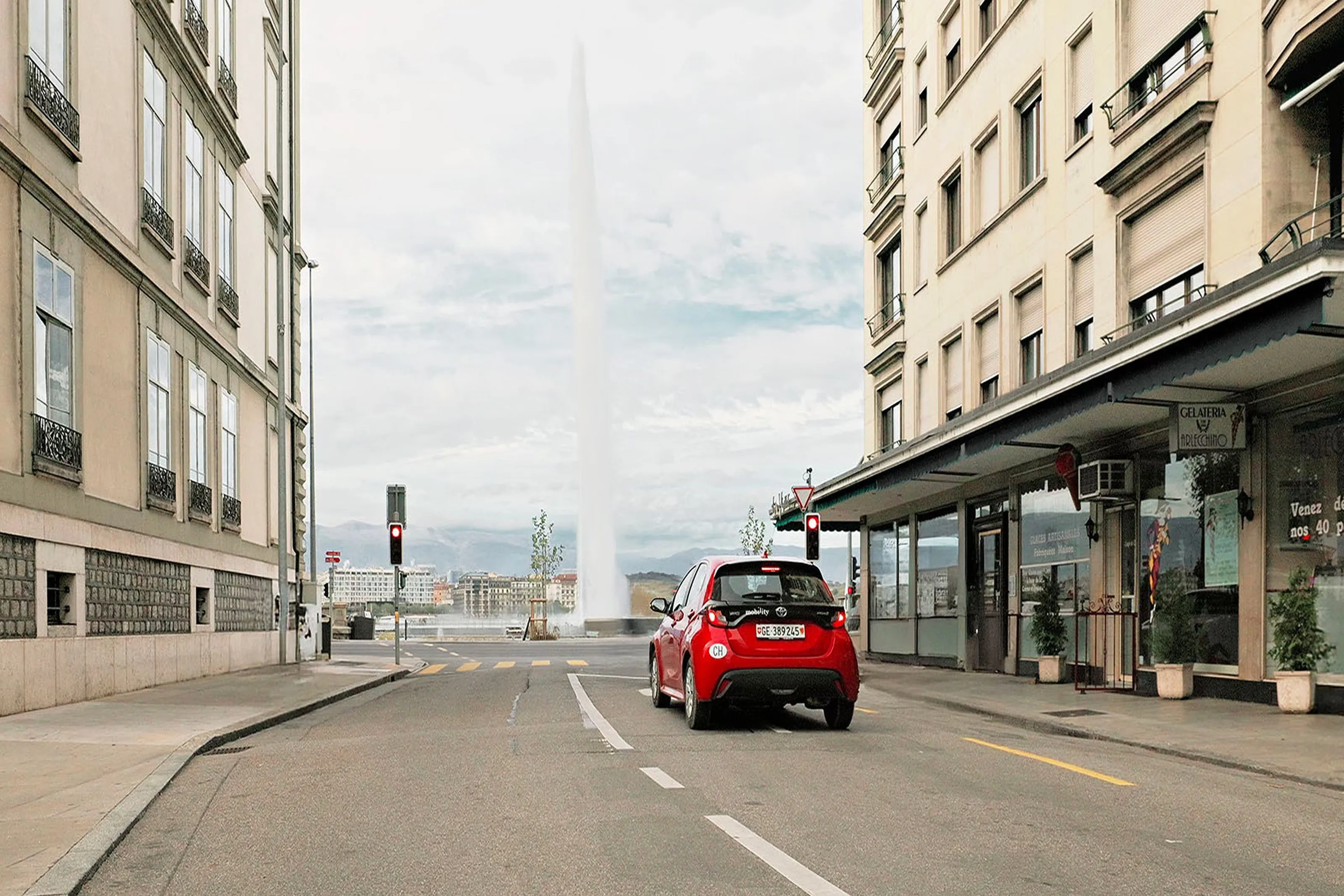 Rotes Mobility-Carsharing-Auto fährt durch eine Stadtstrasse in Richtung Genfer Jet-d’Eau-Brunnen.